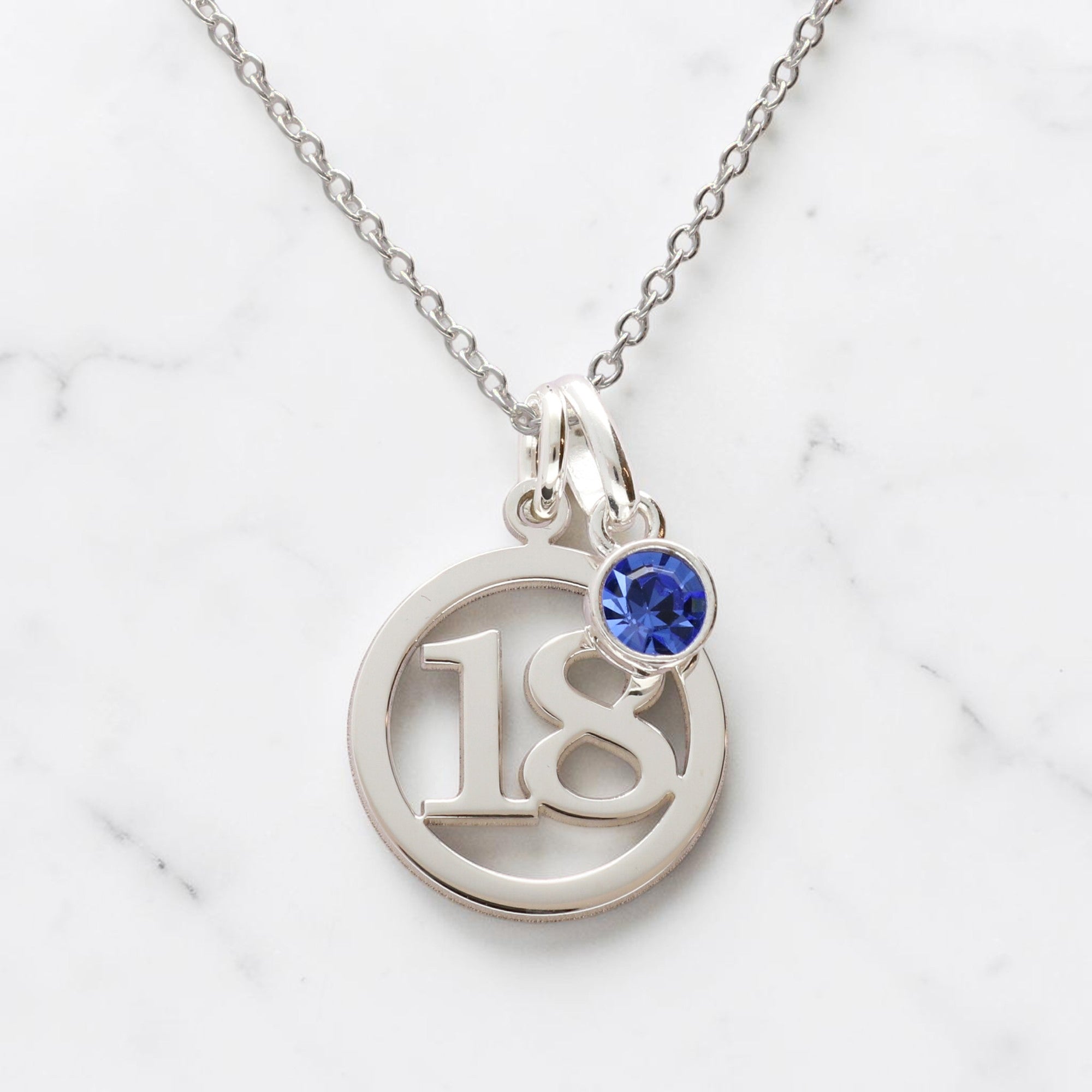 A silver birthday number necklace featuring a circular pendant with the number 18 and a small round blue birthstone stone, displayed on a white marble background.