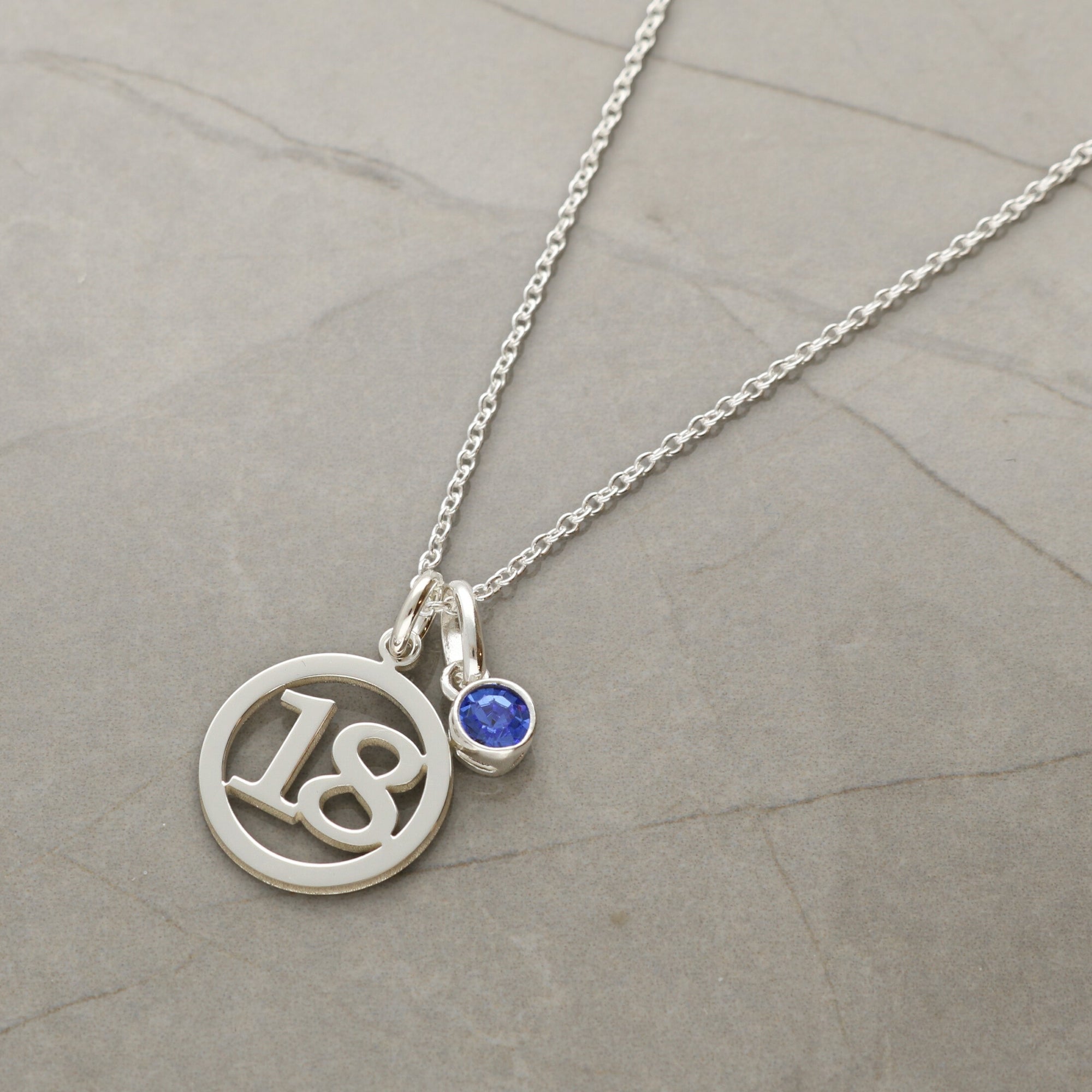 A silver necklace chain with a circular pendant with the number 18 cut out and a small round blue birthstone charm, displayed on a textured gray stone surface.