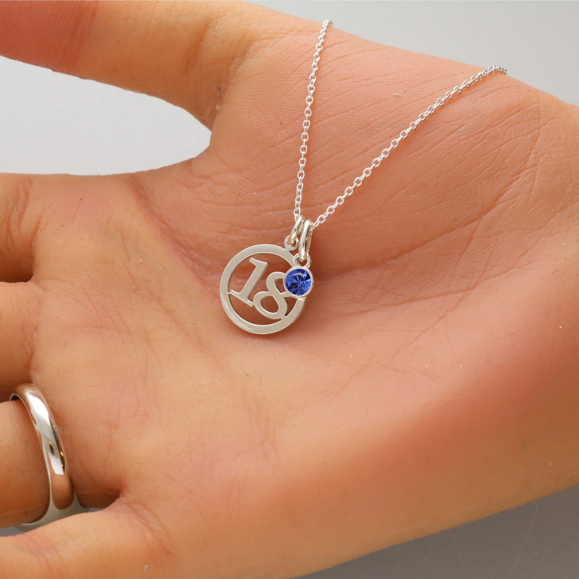A silver pendant necklace with a circular cutout design featuring the number 18 and a small round blue birthstone crystal, displayed on an open hand.
