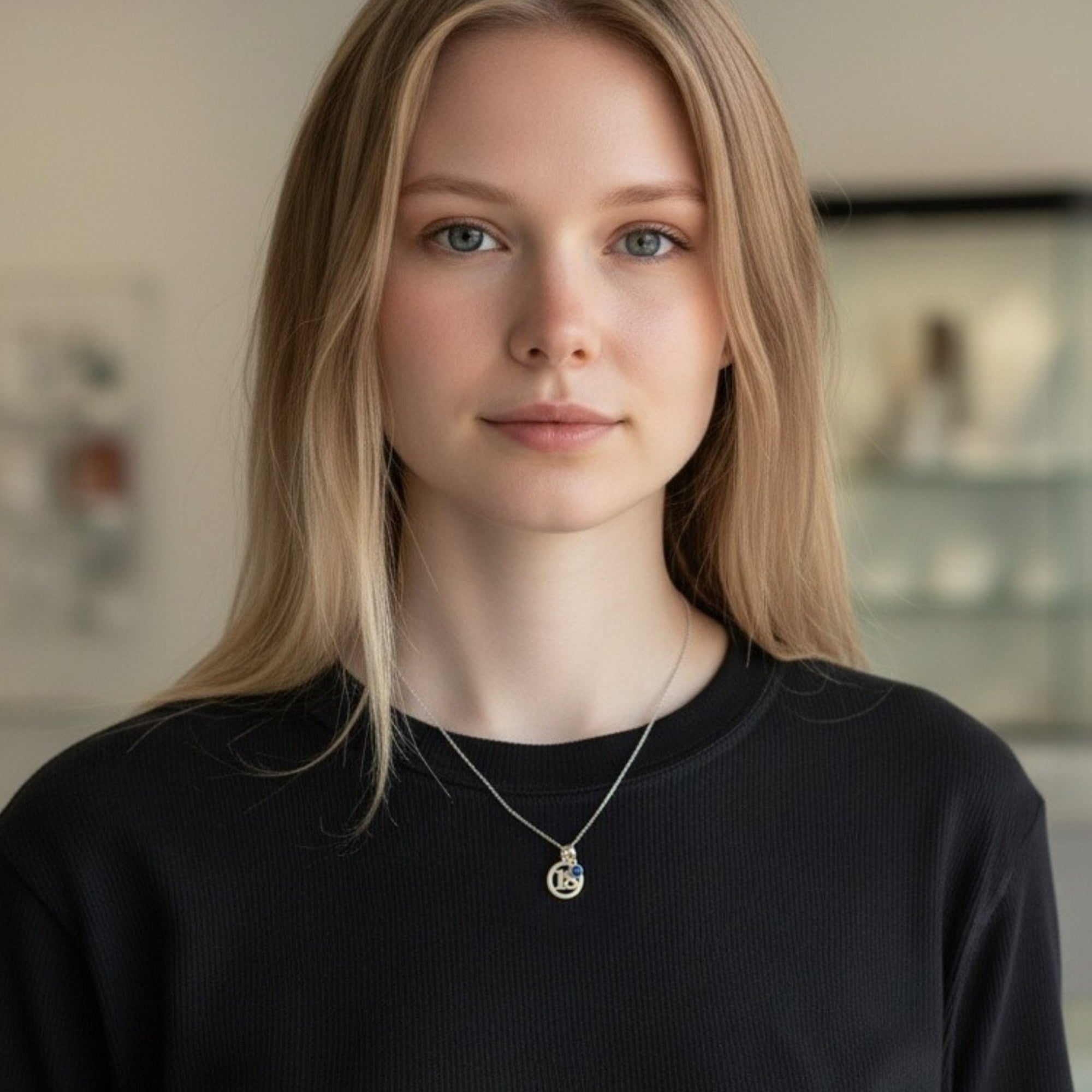 A close-up of a young woman wearing  a silver 18 number in a circle pendant necklace paired with a small circular blue birthstone charm.