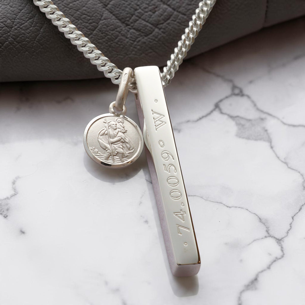 A silver necklace with a round small St Christopher medallion charm and a vertical bar pendant engraved with place coordinates.