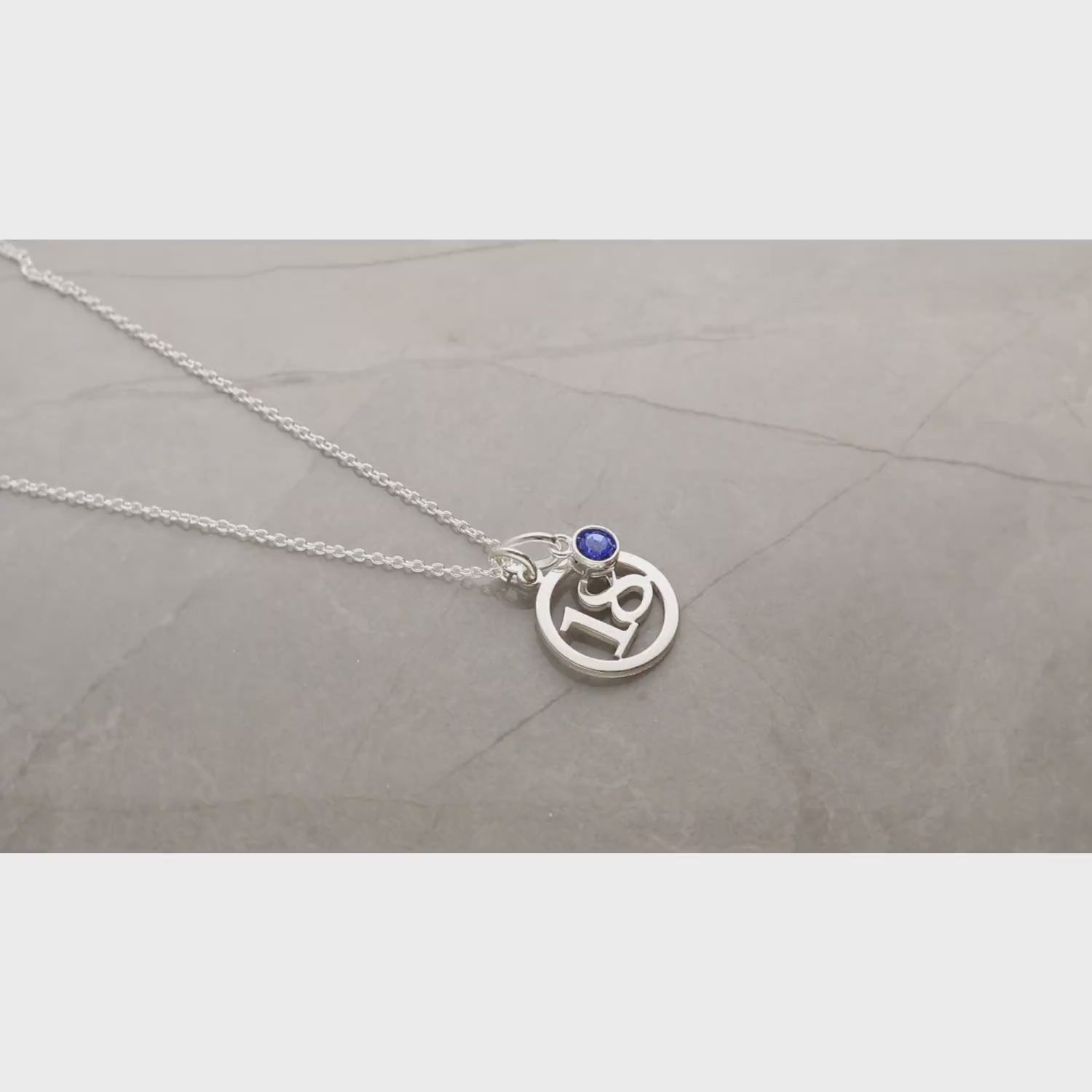 video of silver round number pendant necklace and biue birtstone charm viewed in a 180 degree sweep